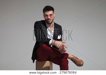 portrait of fashion man wearing grena pants and black suit sitting on wooden box on grey background and fixing his rings