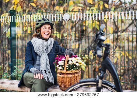 A Senior Woman With Electrobike And Flowers Sitting On A Bench Outdoors In Town.