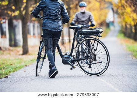 Active Senior Couple With Electrobikes Standing Outdoors On A Road In Park.