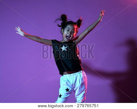 Joyfull Asian Kid Girl In Shirt And Pants With Stars On Purple Background Throws Hands Up Like A Win