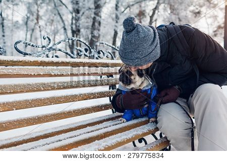 Pug Dog Sitting On Bench With His Master. Puppy Wearing Winter Coat. Man Hugging His Pet In Winter P