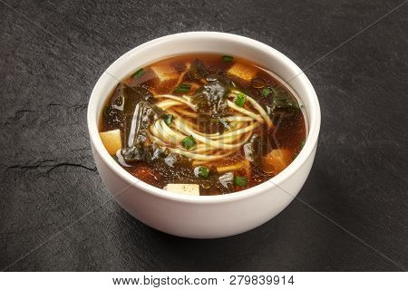 A Photo Of A Bowl Of Miso Shiru Soup With Tofu, Scallions, Noodles, And Wakame Seaweed, On A Black B