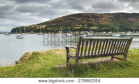 Close-up bench on the Arran island shore. Amazing bayview. Breathtaking Scottish landscape. Cozy bay with the yachts under the rainy cloudy sky. Ideal place for the rest and relax.