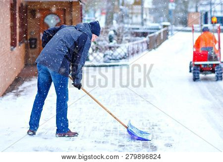 Man With Snow Shovel Cleans Sidewalks In Winter During Snowfall. Winter Time In Europe. Young Man In