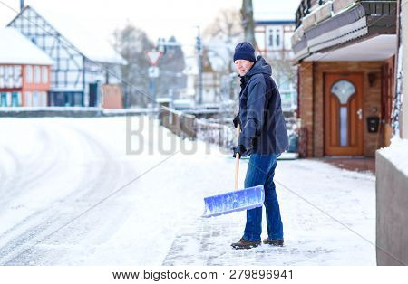 Man With Snow Shovel Cleans Sidewalks In Winter During Snowfall. Winter Time In Europe. Young Man In