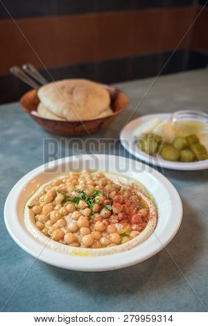 Having an Israeli lunch at cafe in Tel Aviv, Israel. Creamy hummus with whole cheakpeas close up, pile of pitas and pickles on the background