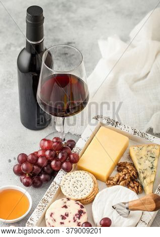 Glass And Bottle Of Red Wine With Selection Of Various Cheese In Wooden Box And Grapes On Light Back