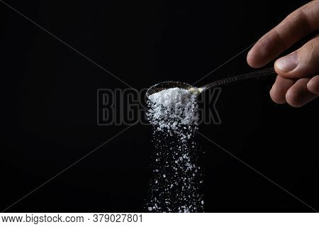 Salt Is Poured From A Spoon On A Black Background. Excessive Salt Intake. Coarse Salt