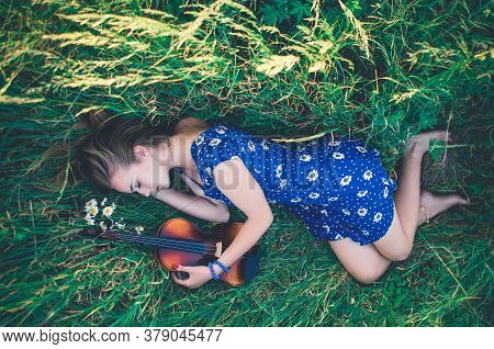 Young Slim Fair-skinned Girl Blonde Beautiful Lies In Tall Grass. A Woman Hugs Violin.