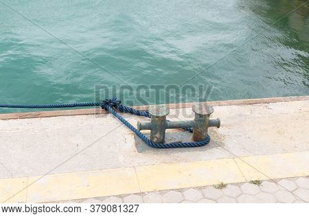 Close-up Of Old Mooring Bollard With A Blue Rope In The Port Against The Background Of The Sea. Knec