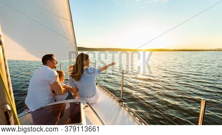 Family Of Three Sailing On Yacht Sitting On Sailboat Deck Looking At Sunset At Seaside. Dream Vacati