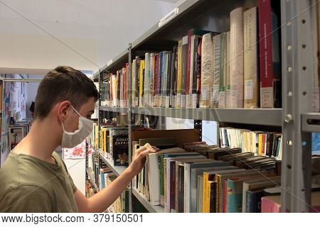 Italy, Summer 2020: A Boy With A Mask For The Covid19 Is Consulting Books In A Public Library. High 