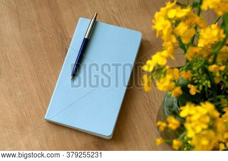 A Blue Notebook With A Pen Lies On A Wooden Table Next To A Vase With Rapeseed Flowers
