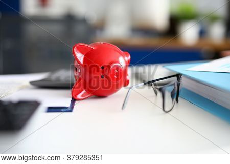 Close-up Of Red Piggy Bank Laying On White Desk. Container For Saving Money. Black Stylish Glasses. 