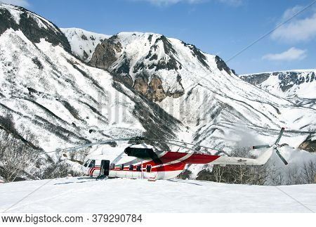 The Helicopter Is Sitting In The Snow In A Mountainous Area. Rotorcraft On A Wild Helicopter Pad In 
