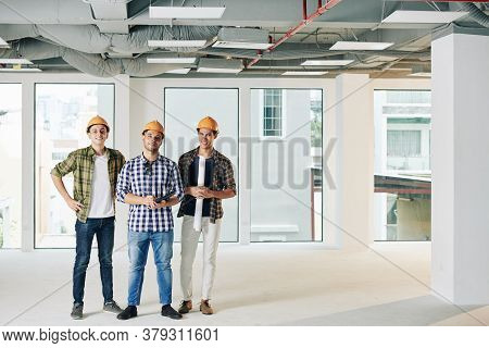 Team Of Smiling Costruction Engineers With Blueprint And Digital Tablet Standing In House Under Cons