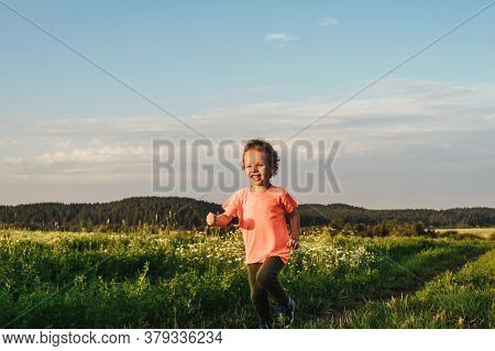 Little Curly Boy In Orange T-shirt Running In A Meadow, Sunset Light. Beautiful Landscape In Summer,