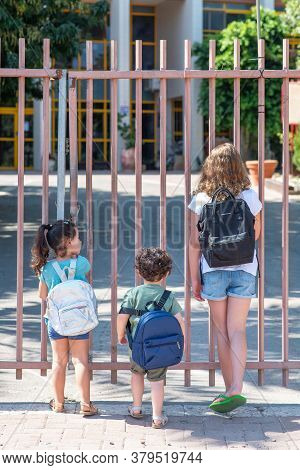 A Young Sisters And Brother Wait Outisde A Closed School Gate. Diverse School Children Going To Scho