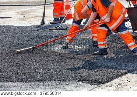 A Working Group Of Road Workers In Orange Overalls Renews A Section Of The Road With Fresh Hot Aspha