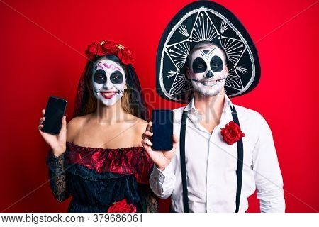 Couple wearing day of the dead costume holding smartphone showing screen looking positive and happy standing and smiling with a confident smile showing teeth 