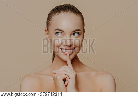 Portrait Of Good Looking Woman Looks Aside Mysteriously, Keeps Finger On Chin And Smiles Broadly, Lo