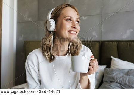 Photo of a young cheerful woman in a bedroom indoors at home listening music with headphones while drinking coffee