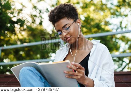 Photo of optimistic african woman sitting outdoors in park while listening music and reading book