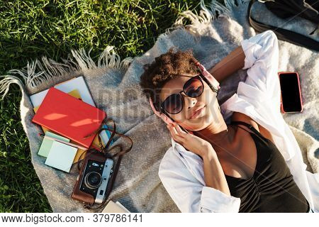 Image of amazing african woman lying outdoors in park on a grass and listening music with heaphones
