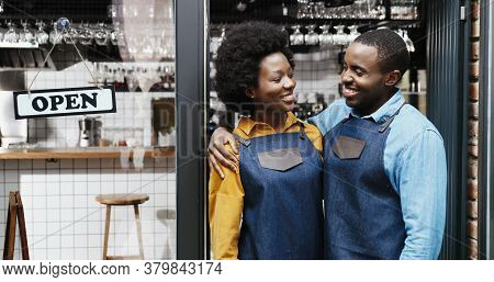 Portrait Of African American Cheerful Couple Of Bartenders In Aprons Hugging Each Other And Smiling 