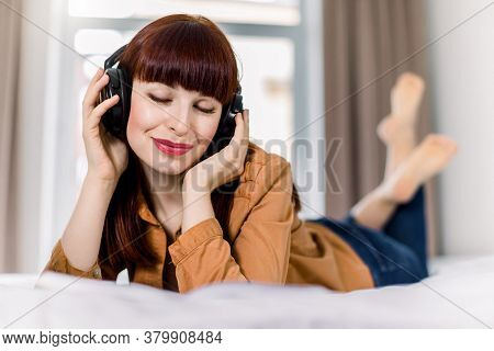 Close Up Of Beautiful Smiling Young Caucasian Woman In Headphones Lying In Bed, With Closed Eyes And