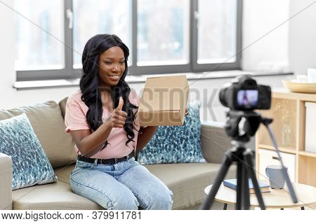 blogging, videoblog and people concept - african american female video blogger with parcel box and camera videoblogging at home
