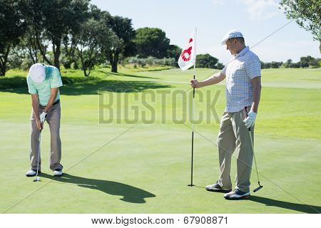 Golfer holding eighteenth hole flag for friend putting ball on a sunny day at the golf course