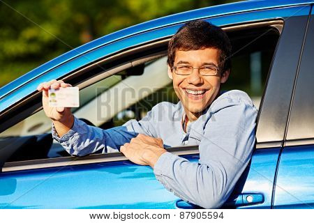 Happy young man in glasses showing his driving license from open car window
