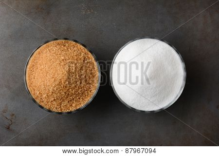 Two bowls of sugar. One bowl of granulated raw turbinado sugar and a second bowl of white granulated sugar. Horizontal format on a used baking sheet.