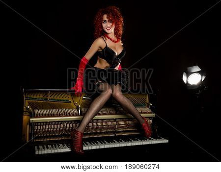 Sexy girl looks like a doll with curly redhead sits on a piano, on a gray background. Fashion style. From the back highlights the soffit