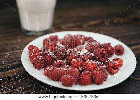 Ripe raspberry on a white plate powdered with sugar. Plate on a wooden table. Sunlight shines through the frame of a window. Glass of yoghurt at the background.