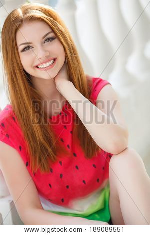 Young beautiful woman with long red, straight hair and green eyes, beautiful make-up, pink plump lips, sweet smile, spending time alone outdoors in the summer in the city, dressed in a red blouse, posing in the afternoon in the sunligh