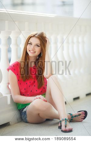 Young beautiful woman with long red, straight hair and green eyes, beautiful make-up, pink plump lips, sweet smile, spending time alone outdoors in the summer in the city, dressed in a red blouse, posing in the afternoon in the sunligh