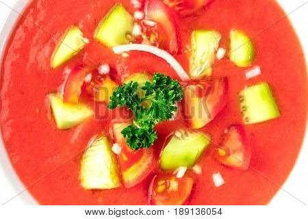 A closeup of gazpacho soup garnished with slices of tomato, cucumber, and onion, with parsley leaves