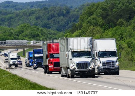 A squadron of eighteen-wheelers lead the way down an interstate highway in eastern Tennessee. Heat waves rising from the pavement give a shimmering effect to vehicles and forest in the background.