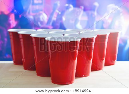 Beer pong tournament layout. Many red party cups in a nightclub full of people dancing on the dance floor in the background. Perfect for marketing and promotion for event. Plastic mugs on wooden table