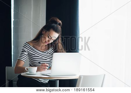 Young woman is sitting at wooden desk near window and making notes. Adult attractive female is sitting with a laptop and a cup of hot coffee while writing in a notebook