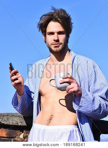 Bearded man short beard. Caucasian macho with moustache and ruffled hair in blue striped pajamas holding cup with cell phone in roof and show strong torso with six packs on blue sunny sky background