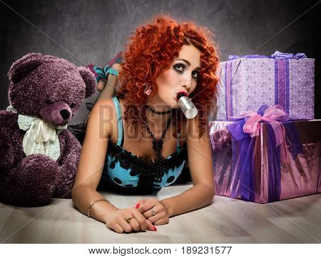 Curly redhead sexy girl looks like a doll for adult men. popsy sucks from a small bottle. Beside sits teddy bear and gift boxes