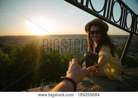 Female giving coffee to man while they sitting on the hill on sunrise city on background