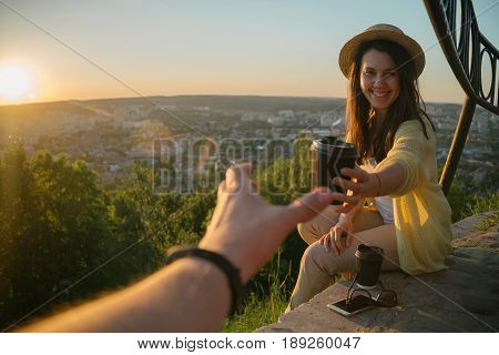 Female giving coffee to man while they sitting on the hill on sunrise city on background