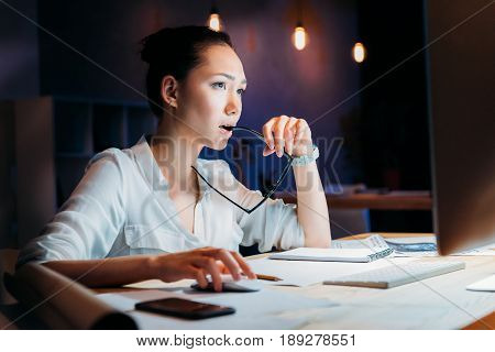 Pensive Asian Businesswoman Holding Eyeglasses And Looking At Computer Monitor, Working Late At The