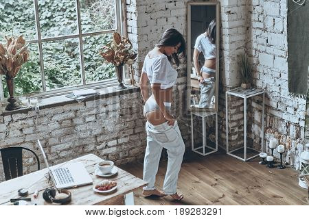 Too sexy for her jeans. Full length rear view of attractive young woman looking in the mirror while putting on her jeans