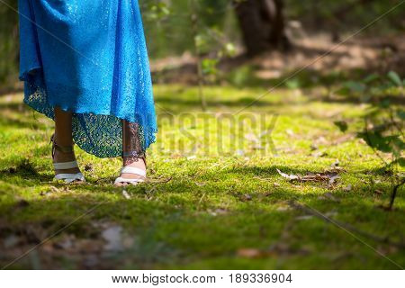 A girl in a blue lace dress and shoes lost her way in the forest. Her feet stand on the moss in the sunshine