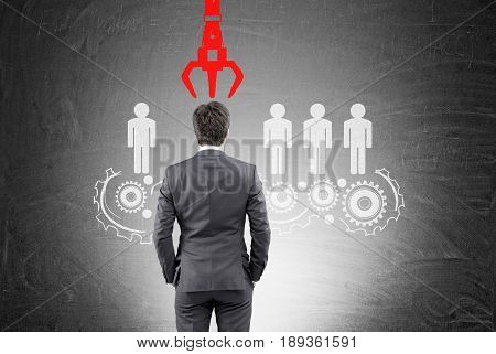 Rear view of a businessman in a suit looking at a white conveyor belt with people figures on it. There is a red grappling hook above his head.